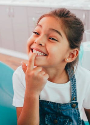 A smiling child wearing a white shirt and denim overalls points at a loose tooth, sitting in a brightly lit indoor space.