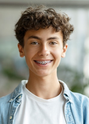 A smiling teenager with braces and curly hair wears a white t-shirt and denim jacket, standing indoors with a blurred background.