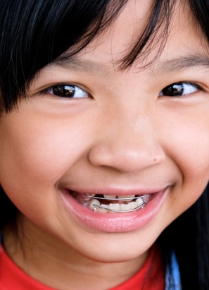 A child with black hair smiles, showing a dental appliance on their teeth. The background is neutral, focusing on the child's face.