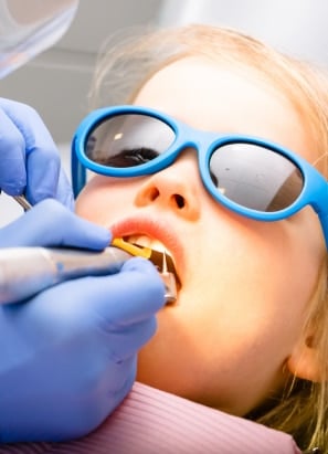 A child wearing blue sunglasses is receiving dental treatment from a gloved person, who is using dental tools in a clinical setting.