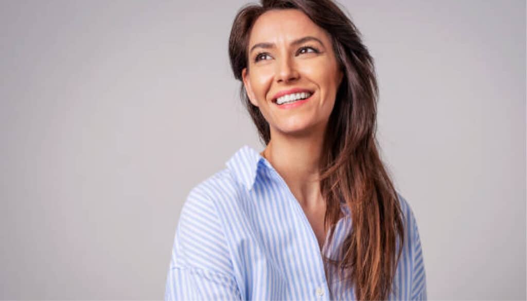 A person with long brown hair and a striped shirt smiles warmly against a plain gray background, with no recognizable landmarks or historical buildings visible.
