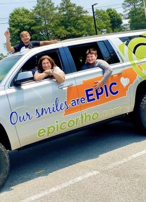 A vehicle with "Our smiles are EPIC" on it has three people smiling from the windows. Trees are visible in the background.
