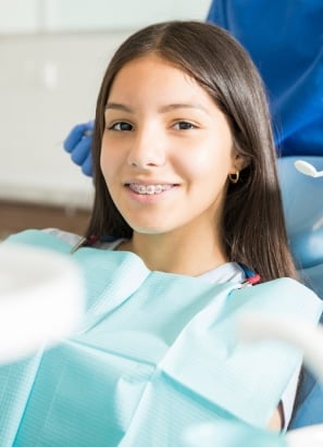 A person sitting in a dental chair, smiling with braces, a person in blue gloves working in the background in a clinical setting.